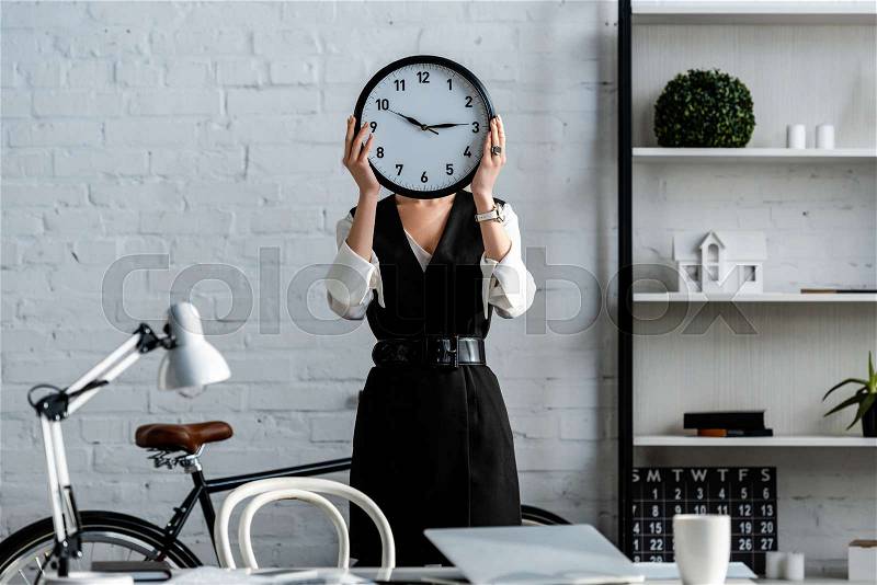 Businesswoman in formal wear holding clock in front of face in office, stock photo
