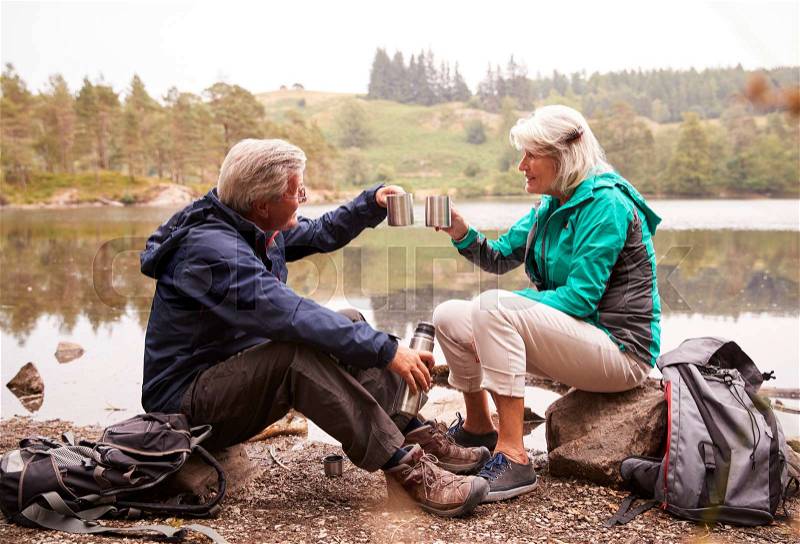 Senior couple sitting by a lake drinking coffee during camping holiday making a toast with their mugs, Lake District, UK, stock photo