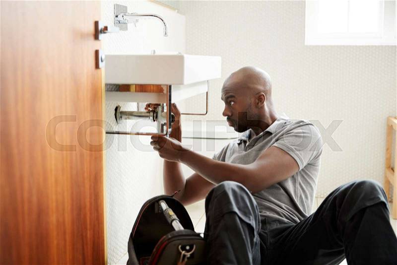 Young black male plumber sitting on the floor fixing a bathroom sink, seen from doorway, stock photo