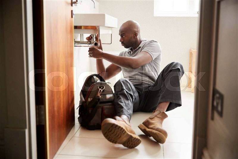 Young black male plumber sitting on the floor fixing a bathroom sink, seen from doorway, full length, stock photo