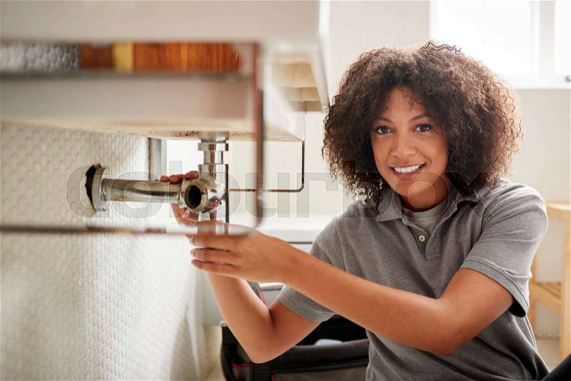 Young black female plumber sitting on the floor fixing a bathroom sink, looking to camera, stock photo
