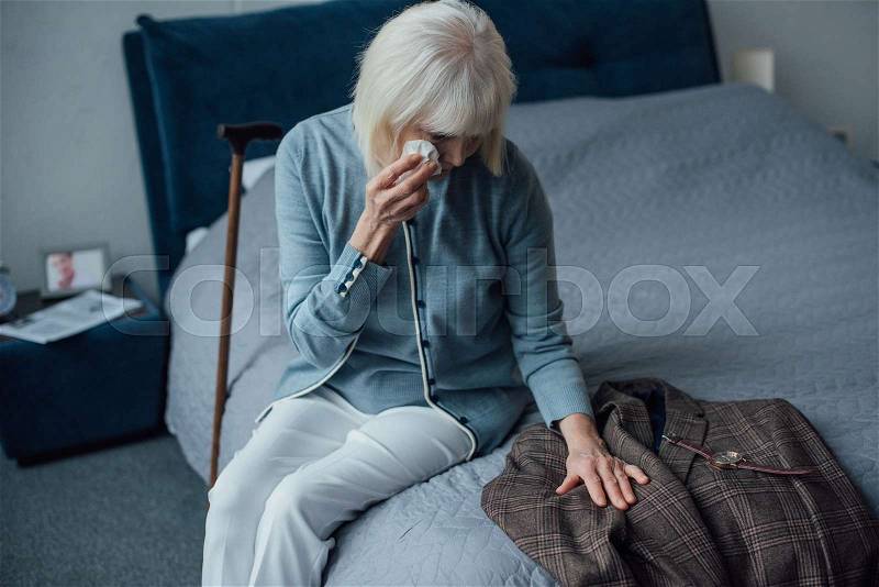 Crying senior woman sitting on bed, wiping tears and looking at jacket at home, stock photo