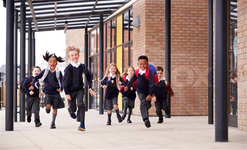 Primary school kids, wearing school uniforms and backpacks, running on a walkway outside their school building, front view, stock photo