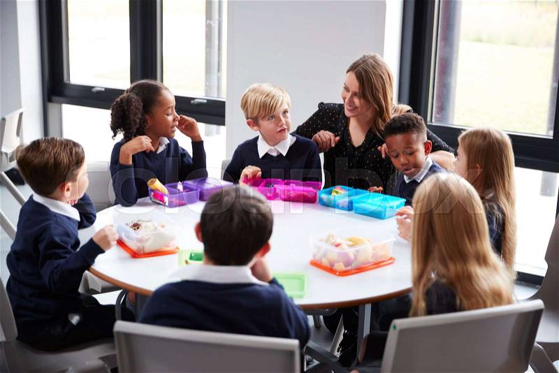Female teacher kneeling to talk to a group of primary school kids sitting together at a round table eating their packed lunches, stock photo