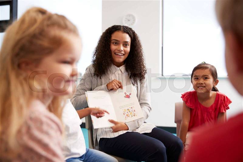Over shoulder view of female teacher showing a picture in a book to a group of kindergarten children sitting on chairs in a classroom, close up, stock photo