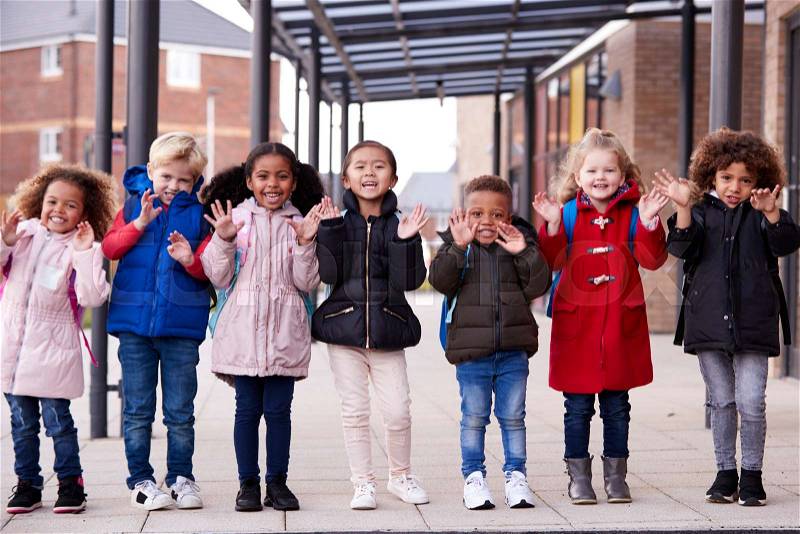 A group of smiling young multi-ethnic school kids wearing coats and carrying schoolbags standing in a row in walkway outside their infant school waving to camera, ..., stock photo