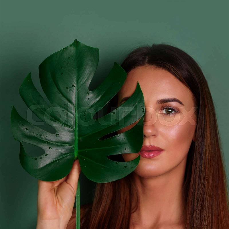 Young woman with natural make up and tropical monstera leaf in hand over green background with copy space. Skin care, pure beauty, cosmetics concept. Square crop, stock photo