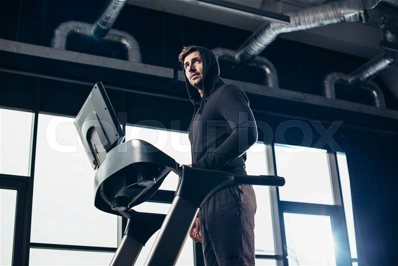 Low angle view of handsome sportsman in hoodie exercising on treadmill in gym, stock photo