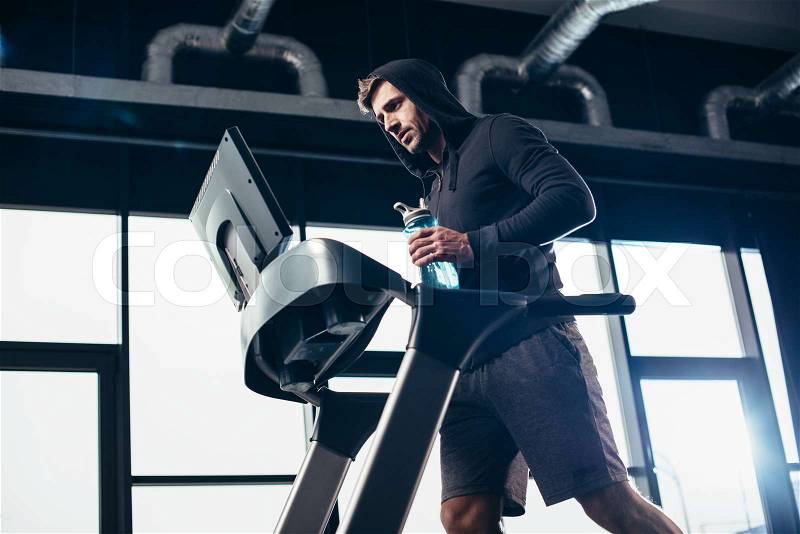 Low angle view of handsome sportsman in hoodie exercising on treadmill and holding sport bottle with water in gym, stock photo