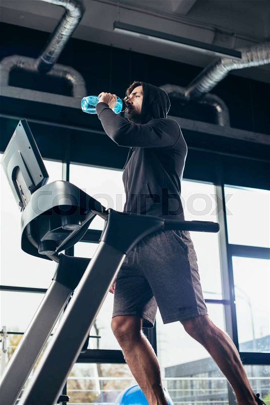 Low angle view of handsome sportsman in hoodie exercising on treadmill and drinking water in gym, stock photo