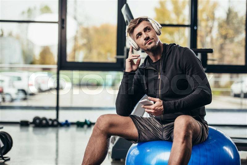 Handsome sportsman sitting on fitness ball and listening music with smartphone in gym , stock photo