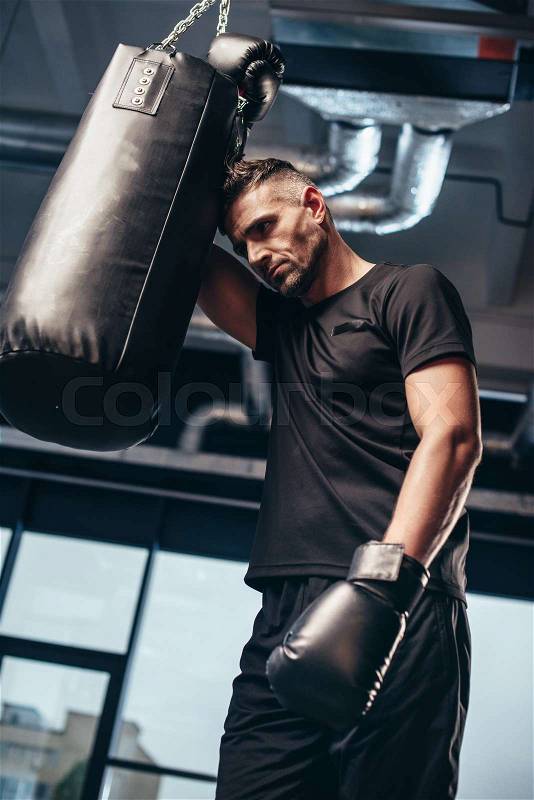 Low angle view of handsome tired boxer leaning on punching bag in gym, stock photo