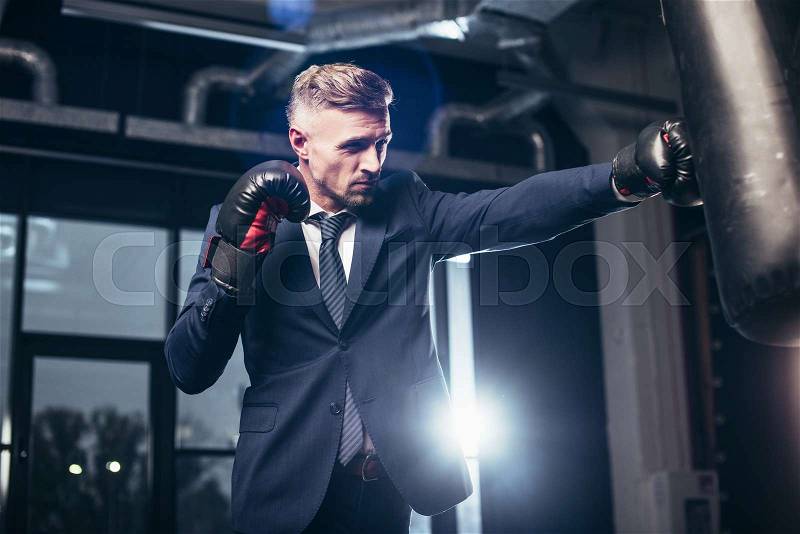 Handsome businessman in suit boxing with punching bag in gym, stock photo