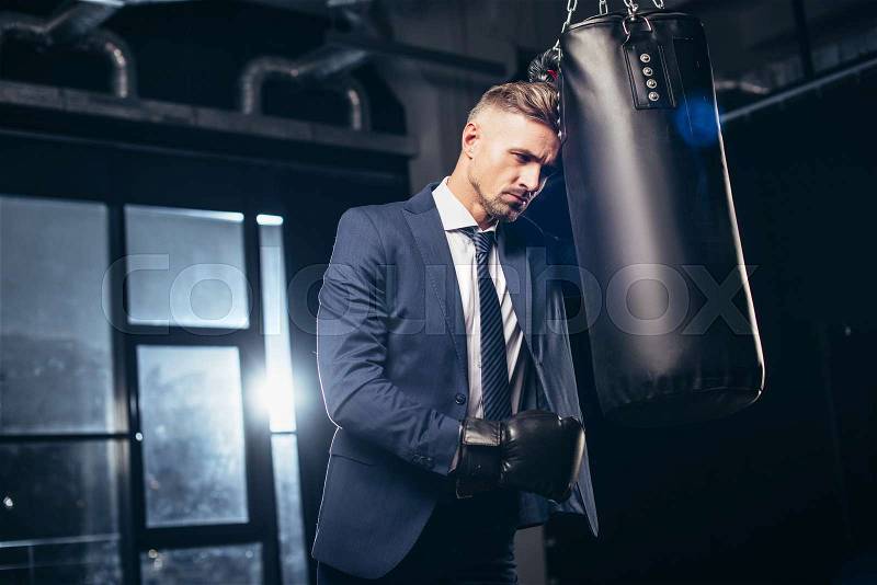 Handsome tired businessman in suit and boxing gloves leaning on punching bag in gym, stock photo