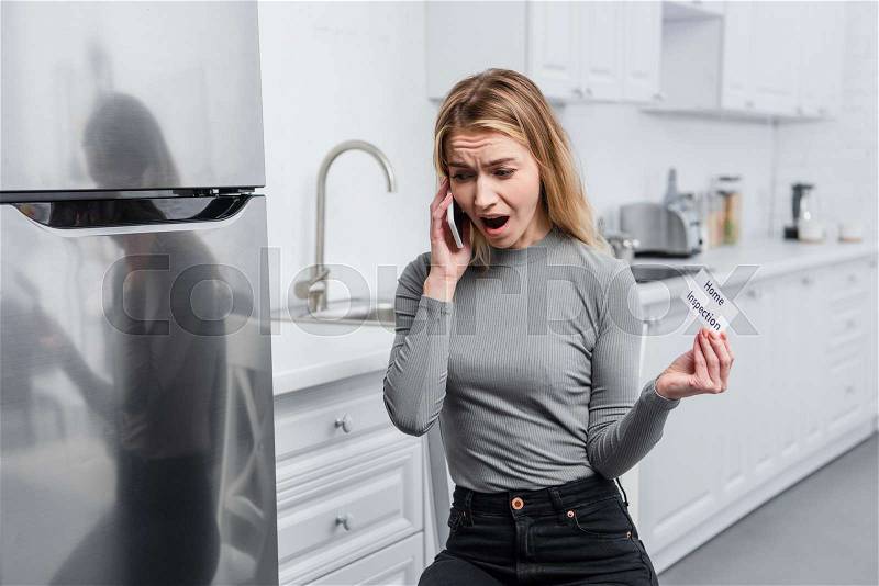 Angry young woman holding card with lettering home inspection and yelling on smartphone in kitchen near broken refrigerator, stock photo