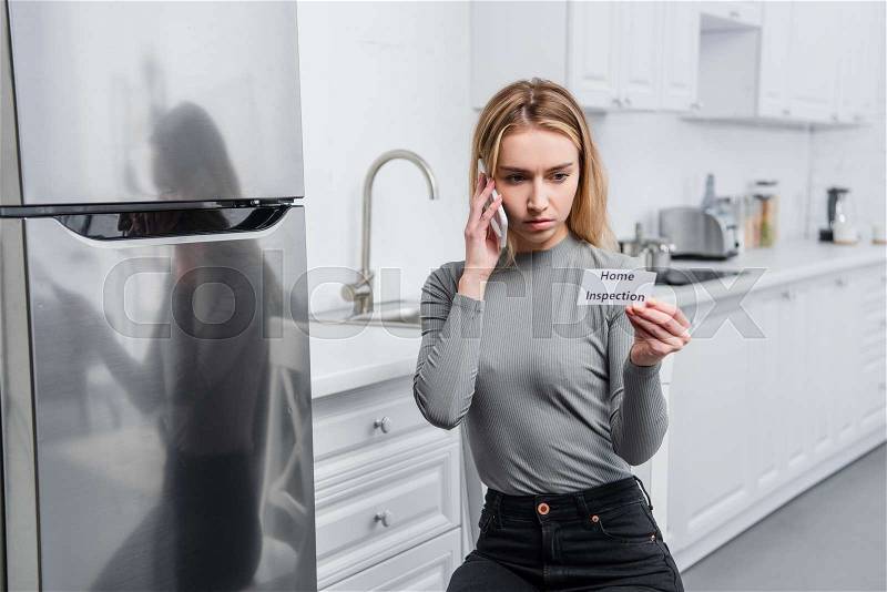 Serious young woman holding card with lettering home inspection and talking on smartphone near broken refrigerator in kitchen , stock photo