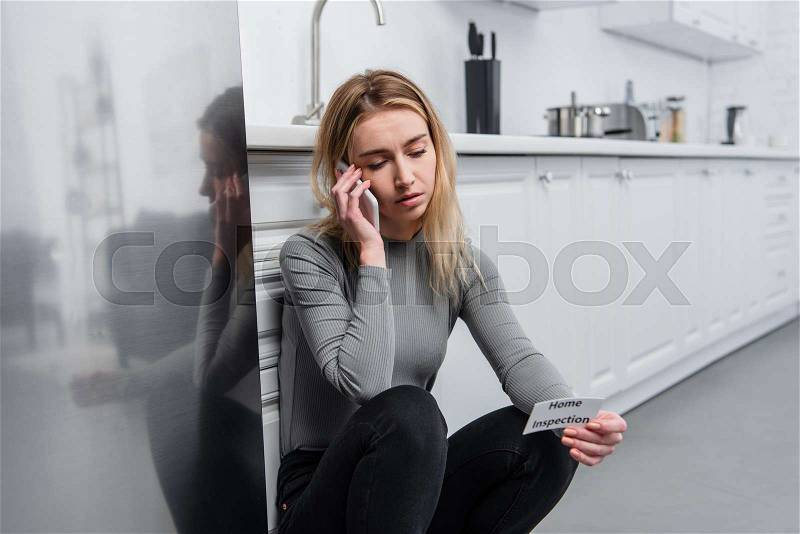 Blonde young woman holding card with lettering home inspection and talking on smartphone in kitchen near broken fridge, stock photo