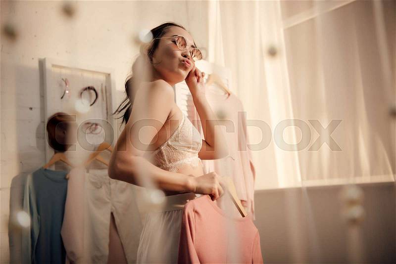Low angle view of beautiful young woman in underwear and sunglasses choosing clothes at home, stock photo