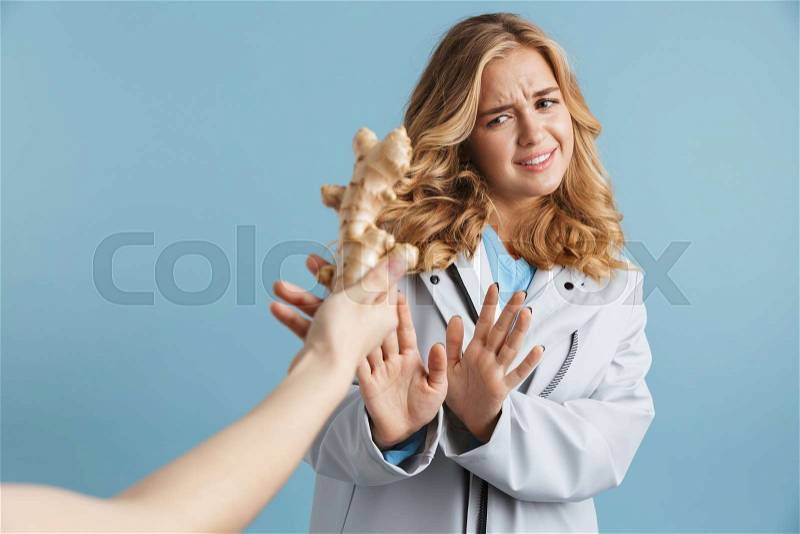 Image of dissatisfied woman 20s wearing raincoat doing stop gesture and looking at ginger root, isolated over blue background, stock photo