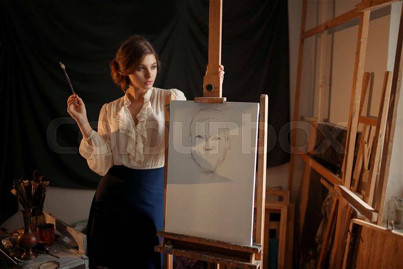Cute female painter with brush standing against easel in studio. Creative paint, woman drawing pencil sketch, workshop interior on background, stock photo