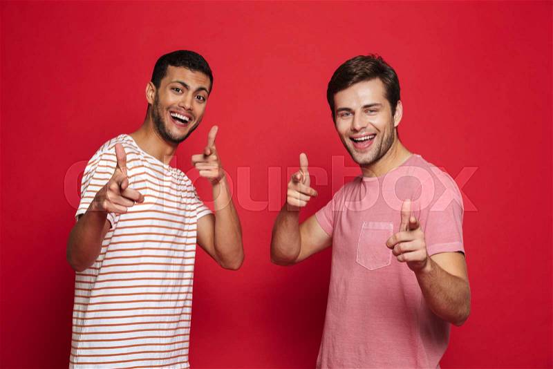 Two cheerful young men standing isolated over red background, pointing at camera, stock photo