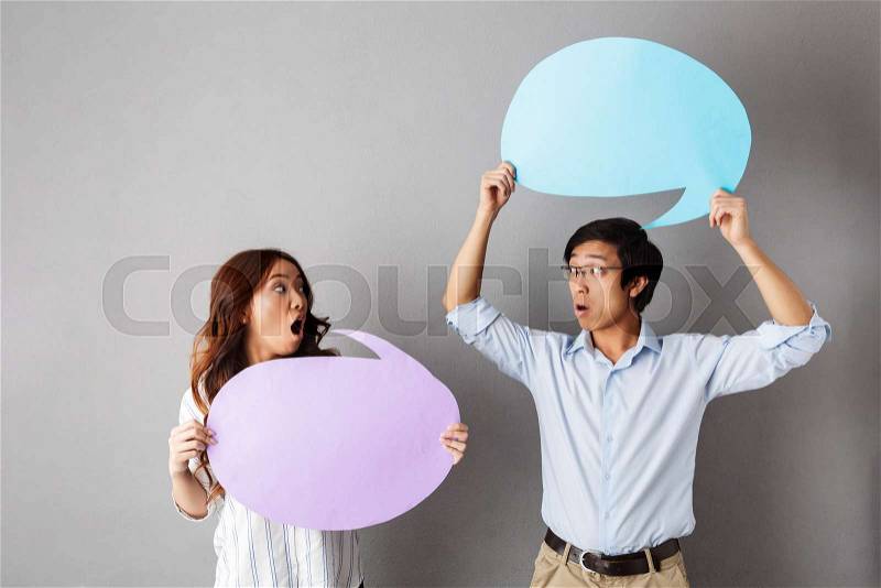 Confused asian couple standing isolated over gray background, holding empty speech bubble, having an argument, stock photo