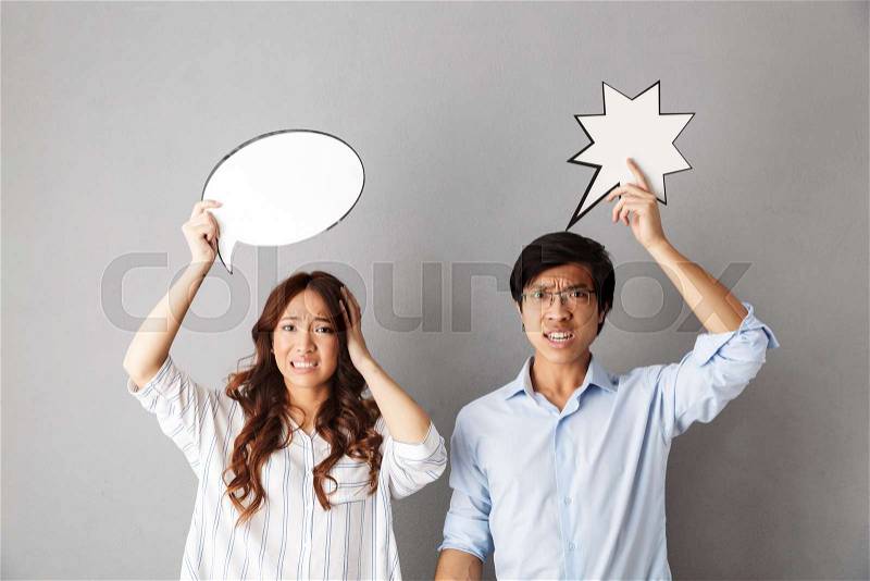 Confused asian couple standing isolated over gray background, holding empty speech bubble, having an argument, stock photo