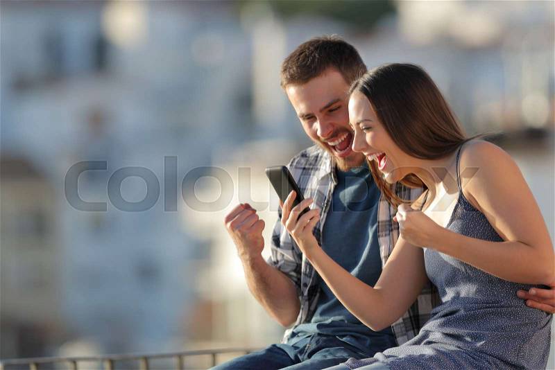 Excited couple celebrating online smart phone news outdoors in a town outskirts at sunset, stock photo