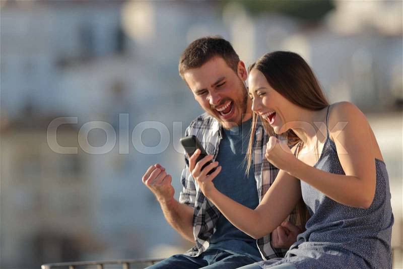Excited couple or friends reading smart phone content in a town at sunset, stock photo