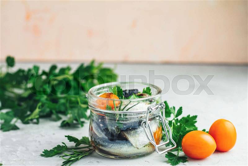Sardines or baltic herring with rosemary, thyme, parsley, tomatoes slices and spaces in glass jar on light gray concrete table surface. Preserved marinated sea fish, ..., stock photo