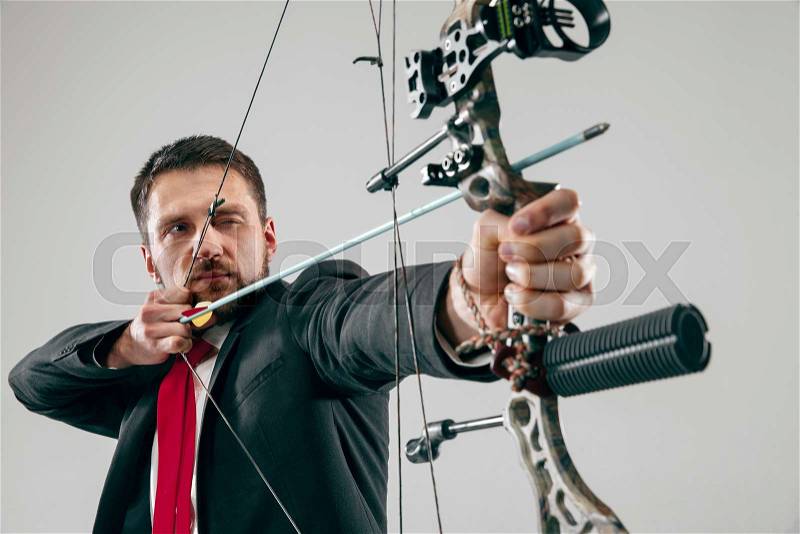 Businessman aiming at target with bow and arrow, isolated on gray studio background. The business, goal, challenge, competition, achievement concept, stock photo