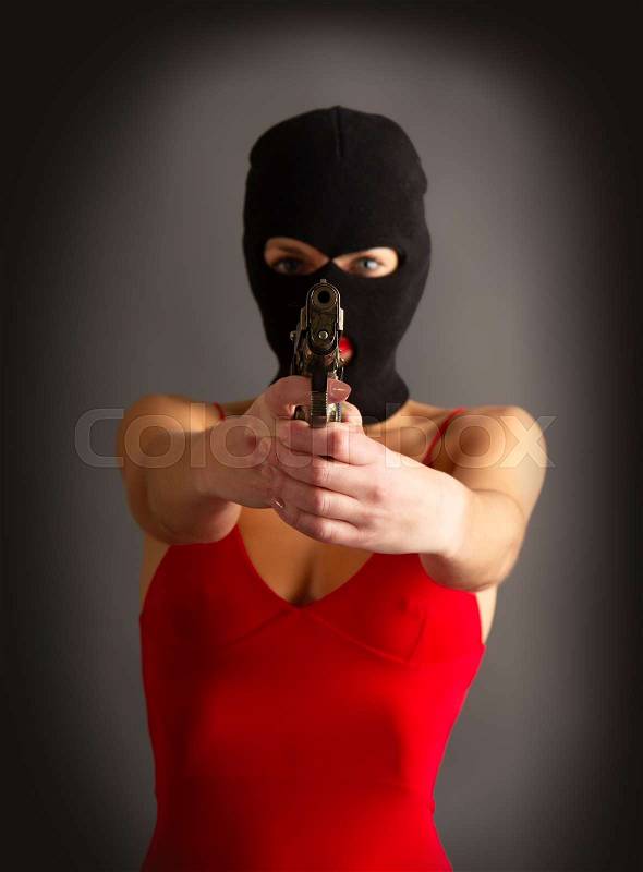 Young self-confident girl with a big gun in a black balaclava and in a red sexy dress standing on a gray background, stock photo