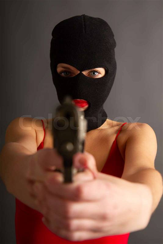 Young self-confident girl with a big gun in a black balaclava and in a red sexy dress standing on a gray background, stock photo