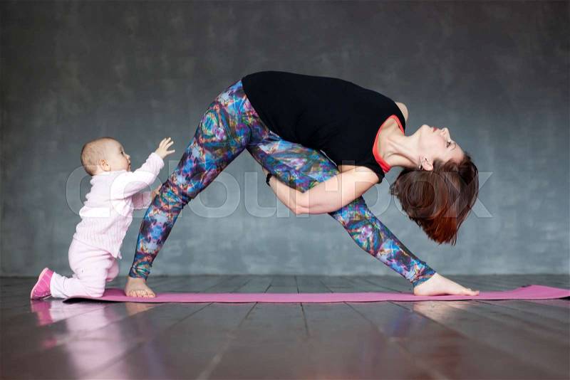 Young woman practicing yoga, standing in Utthita Trikonasana exercise, extended triangle pose, working out. Her pretty baby standing near helping her, stock photo