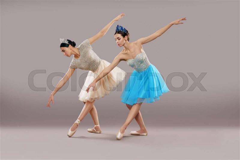 Side view of two professional dancers in dresses, ballet shoes and crowns dancing and bending down in studio. Slim ballerinas performing on grey isolated background. ..., stock photo