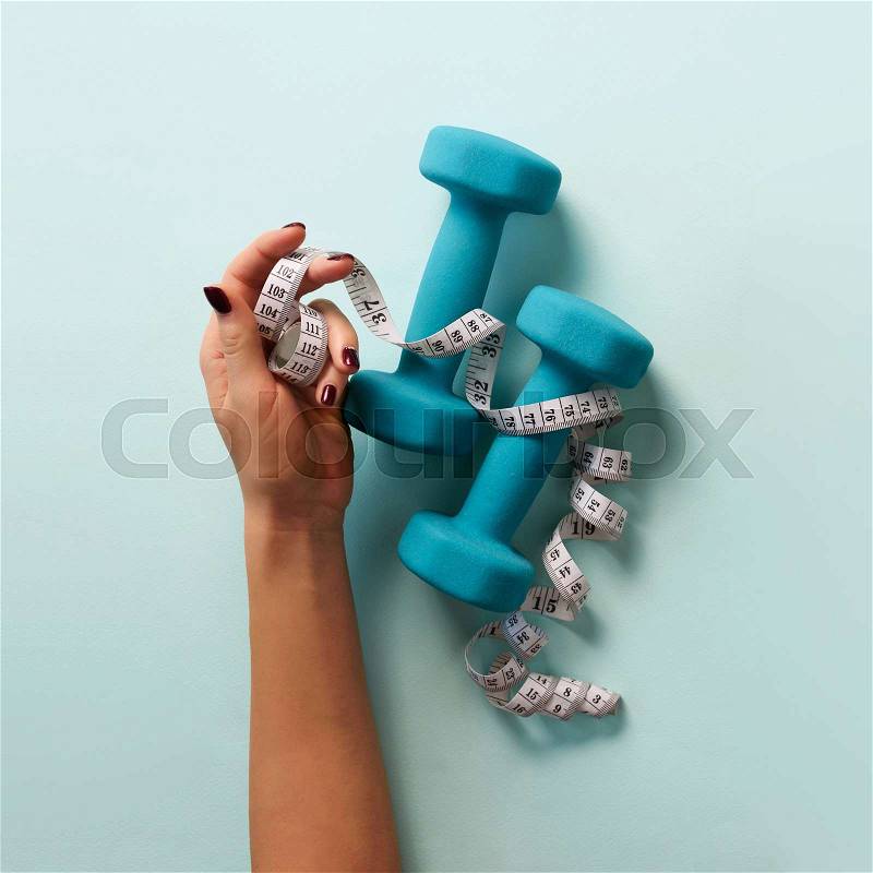 Female hand holding sweet donut, measuring tape, dumbbells over blue background. Top view, flat lay. Weight lost, sport, fitness, diet concept. Banner with copy ..., stock photo
