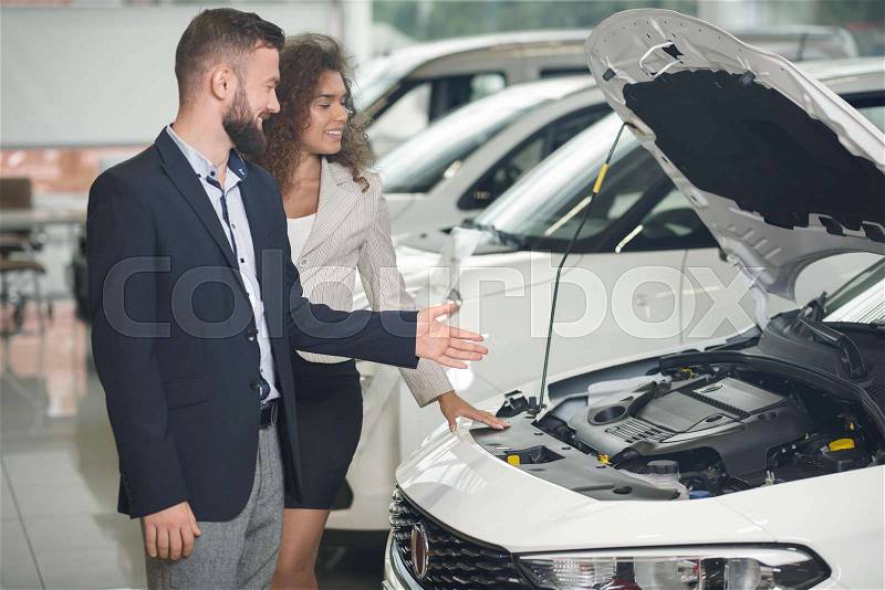 Car dealer showing to female client white automobile in auto showroom. Manager and beautiful woman observing together car under hood. Handsome bearded man wearing in ..., stock photo