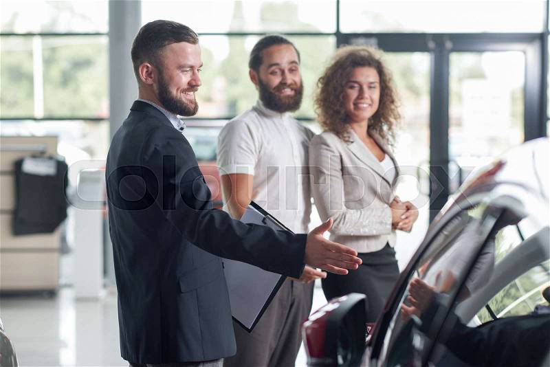Happy clients standing near car dealer and smiling. Men and pretty woman looking at automobile, observing. Manager in dark blue jacket holding folder, showing ..., stock photo
