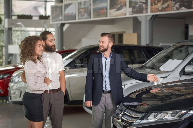 Manager of car dealership showing dark blue auto to clients. Happy couple observing expensive automobiles in car center. Brutal man in white shirt holding hand round ..., stock photo