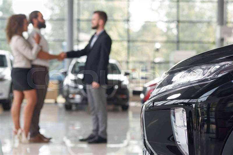 Close up of black car headlight. Couple making deal with car dealer behind. Man and manager of car showroom shaking hands, standing opposite. Purchase of vehicle, stock photo