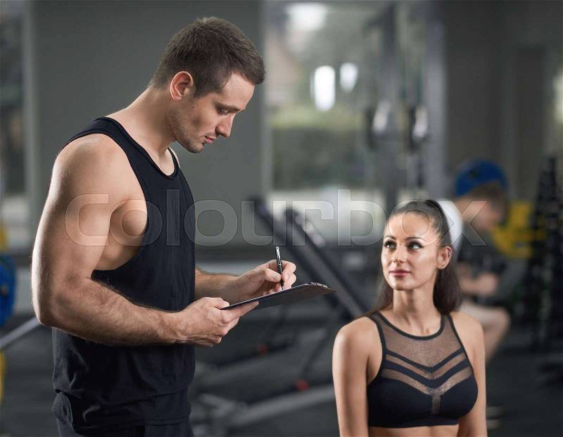 Two sportsmen talking in gym. Professional young athlete helping tan attractive woman become fit and training. Seductive man in black sportswear talking with girl ..., stock photo