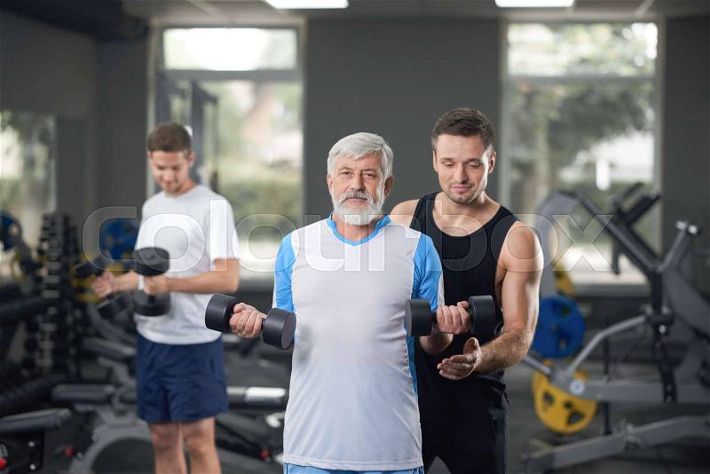 Fit and healthy old man working out with personal trainer in gym. Elderly man lifting dumbbells in hands, looking at camera and posing. Man with gray hair and beard ..., stock photo