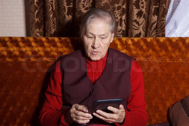 Old woman reading ebook on digital tablet device sitting in her sofa, stock photo