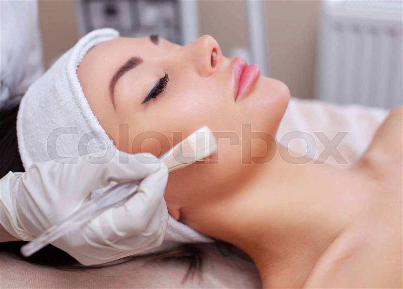 The beautician conducts face cleansing of a beautiful young woman with a soft brush dipped in a moisturizer at a beauty salon. Cosmetology and professional skin ..., stock photo