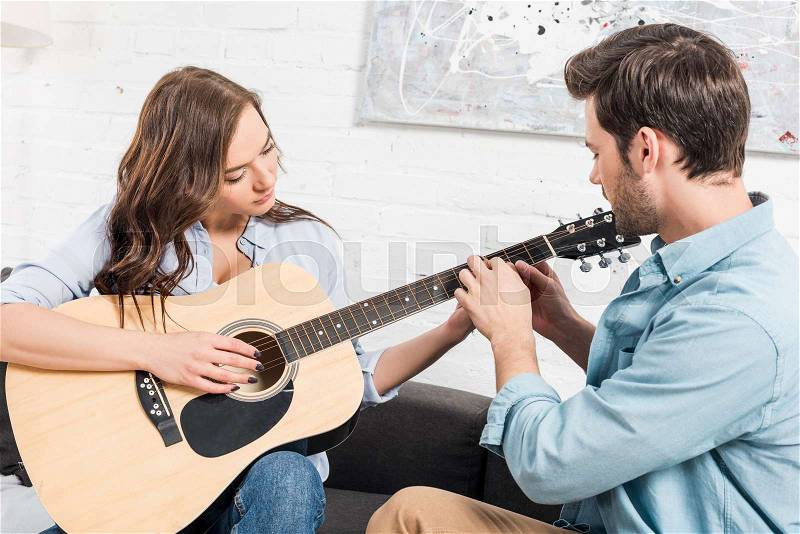 Man sitting on couch and teaching woman to play acoustic guitar at home, stock photo