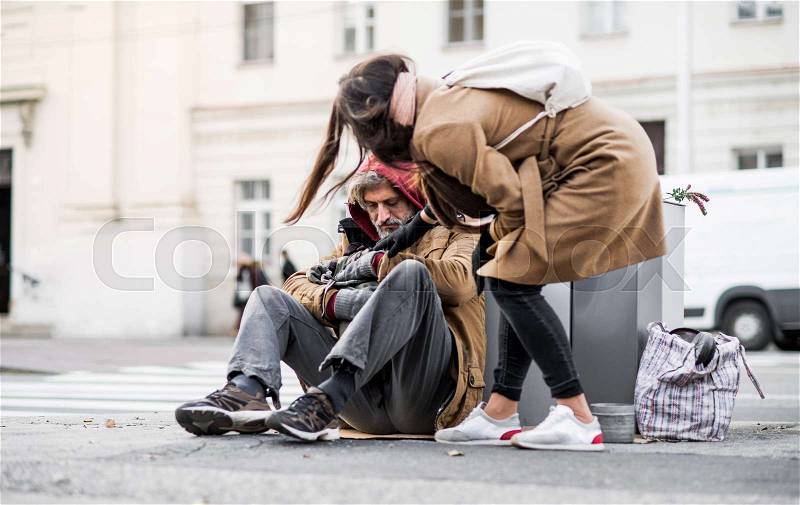 A young woman giving money to homeless beggar man sitting outdoors in city, stock photo