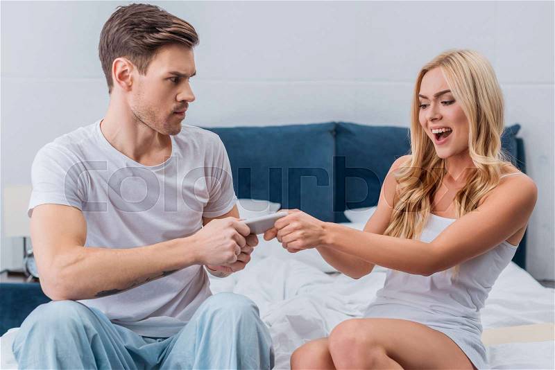 Jealous young woman and unhappy man holding smartphone together on bed, stock photo