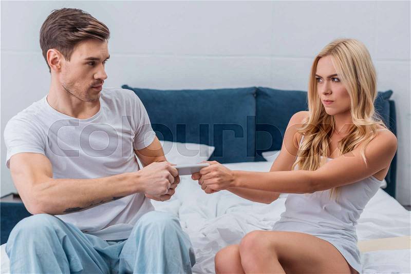 Angry young couple fighting for smartphone on bed, mistrust concept , stock photo