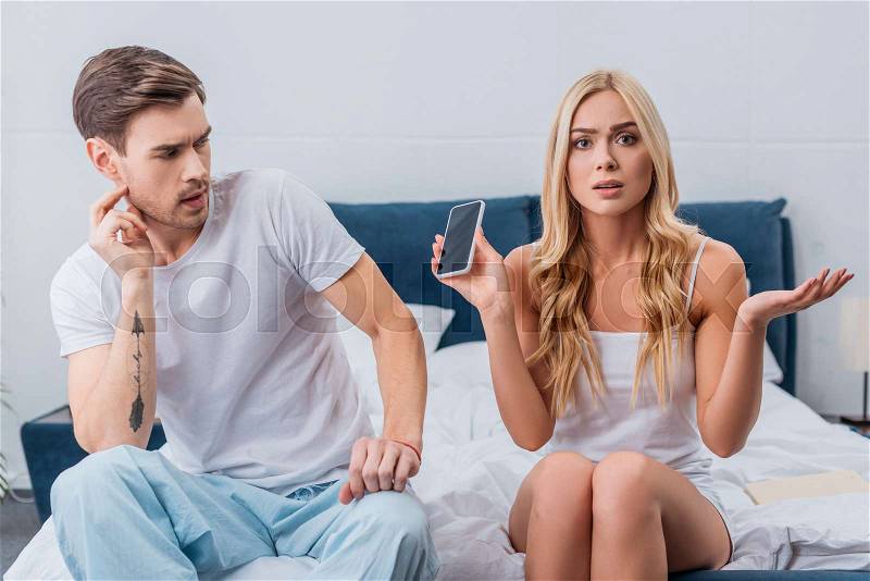 Confused woman holding smartphone and looking at camera while sitting with boyfriend on bed, stock photo