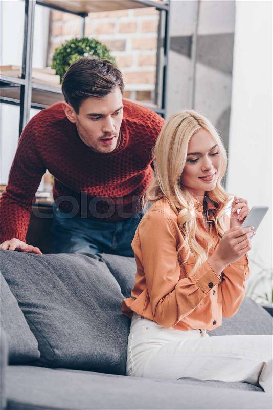 Jealous emotional man looking at smiling young wife using smartphone at home, mistrust concept, stock photo
